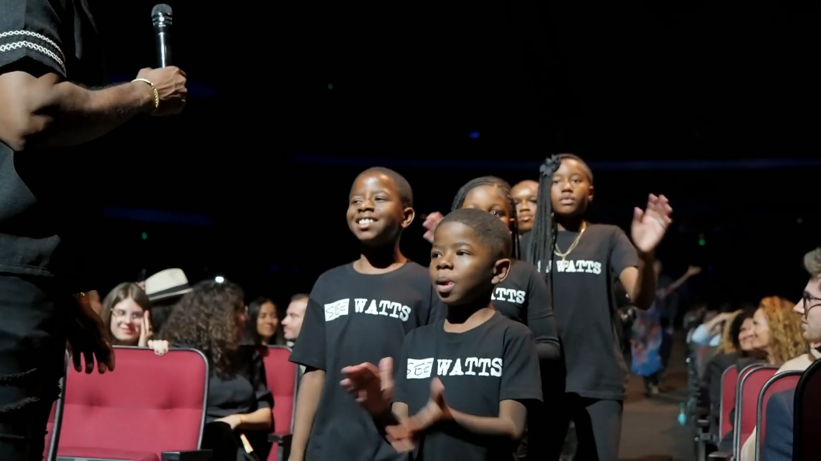 Children in Watts shirts singing in the aisle of the Peacock Theater, the audience around them